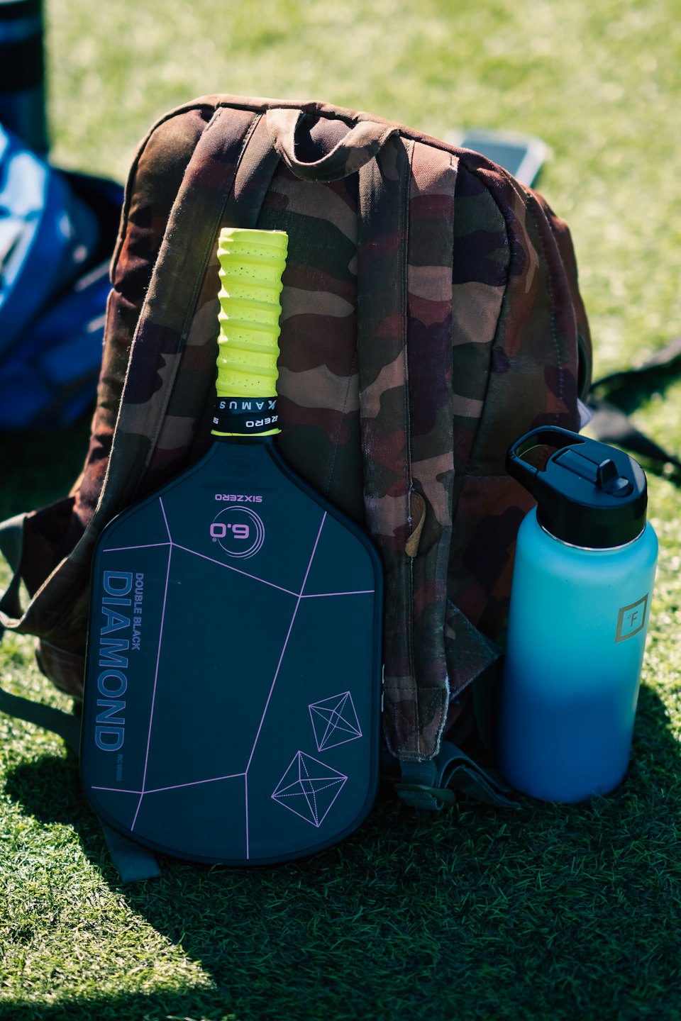 Pickleballracket, ryggsäck och vattenflaska på grönt gräs.